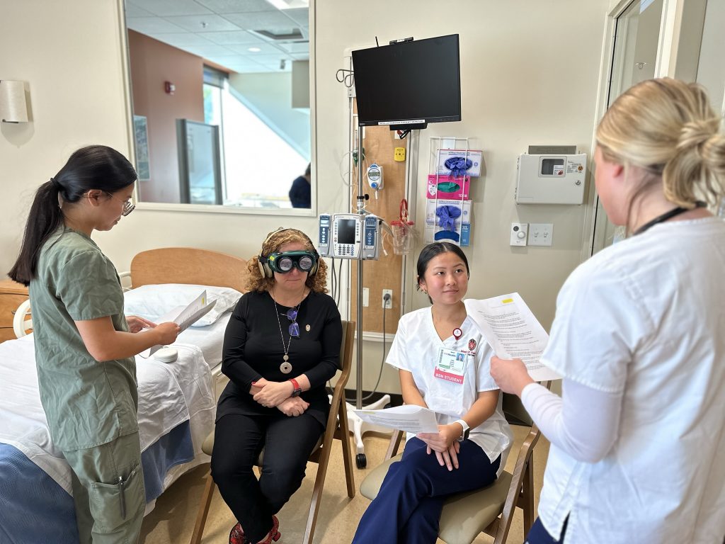 Three students read scripts as part of a simulation scenario. Chancellor Jennifer Mnookin wears impairment goggles and headphones to simulate being a patient with dementia and reduced sensory perception.