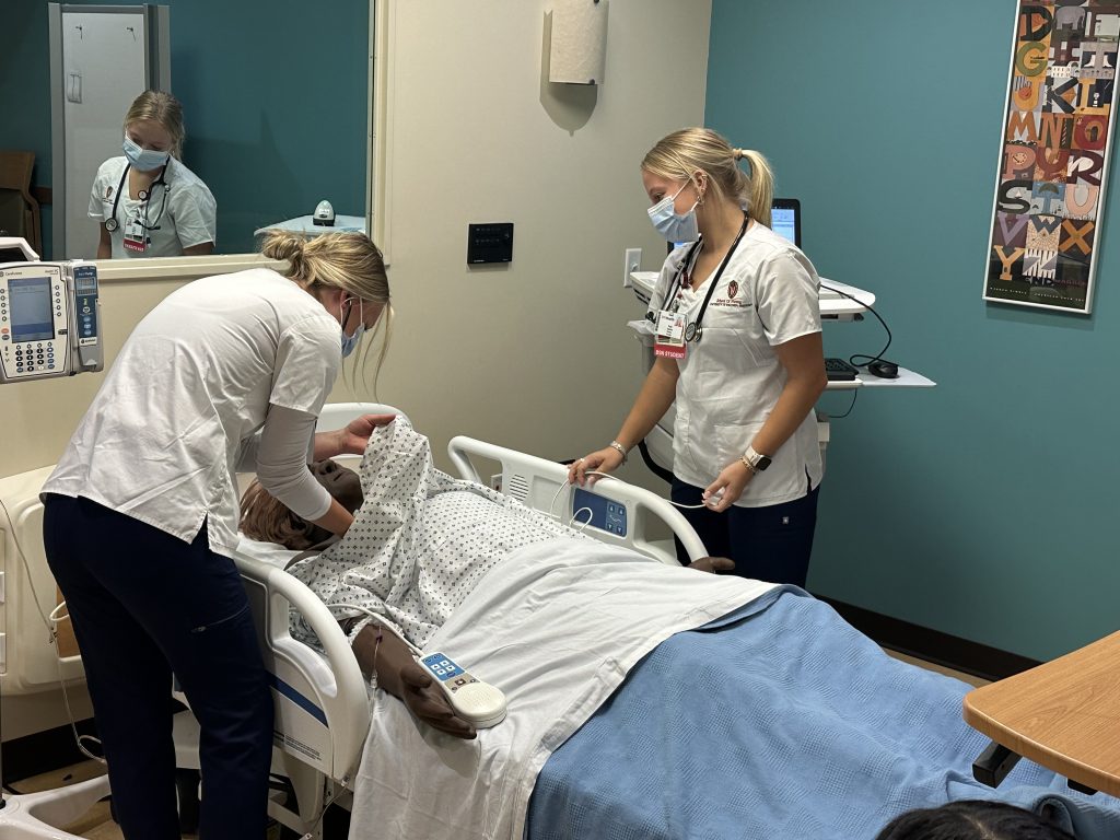 Two female students practice assessment skills on manikin in hospital simulation suite.