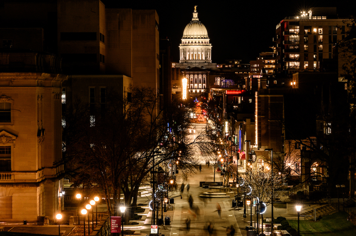 With the Wisconsin state capitol in the background, pedestrians and students walk along Library Mall and the State Street Mall at the University of Wisconsin-Madison after sunset.