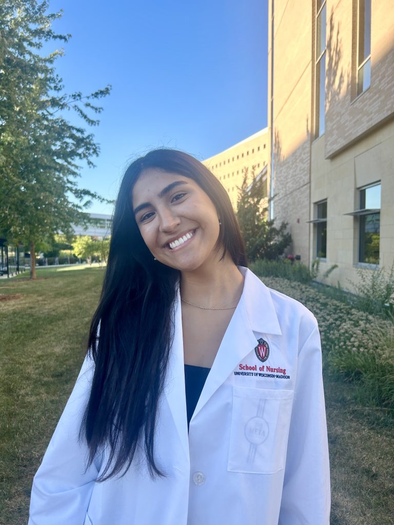 Rachelle Navarro stands outside smiling and wearing her white School of Nursing coat.