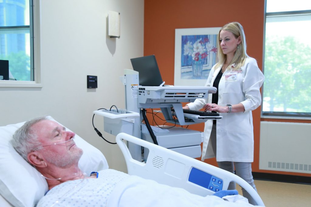 A DNP student looks at information on a computer as a patient lies in a hospital bed.