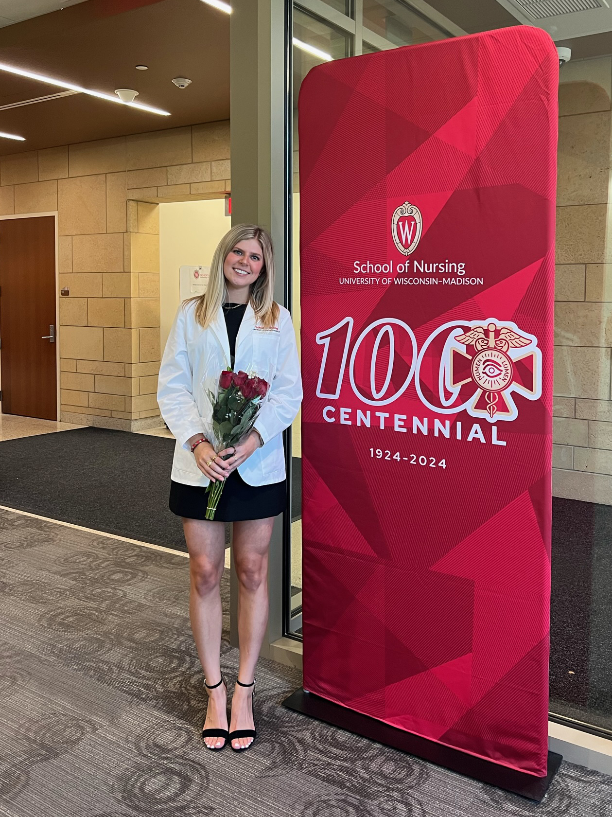 Allie Young holds a bouquet of red roses and poses next to a red School of Nursing Centennial pop-up banner. She wears a black dress and a nursing white coat.