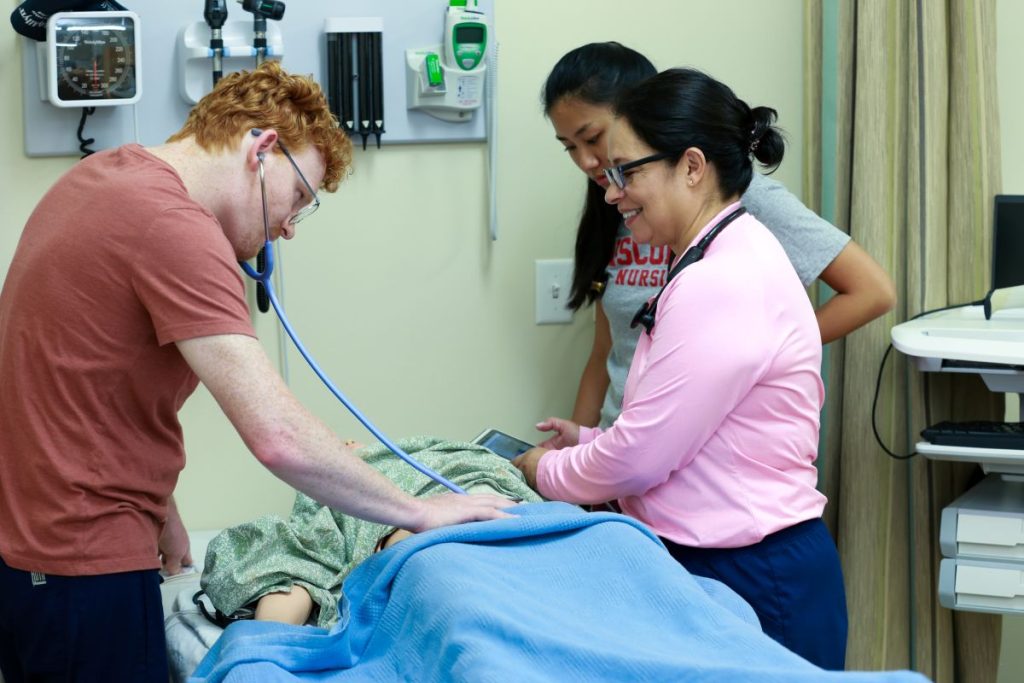 A clinical instructor guides two UW-Madison accelerated nursing program students in examining a medical mannequin.