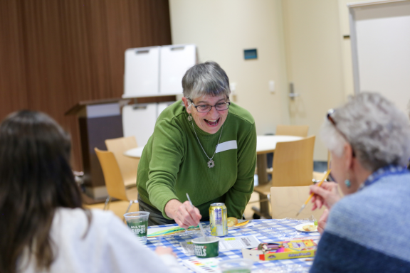 Participant bends down smiling to dip her paintbrush into the paint and continue her painting representing aging.