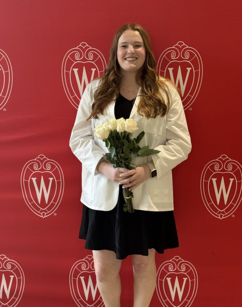 Meghan Debot at the White Coat ceremony, standing against a red UW backdrop holding a bouquet of flowers and smiling.