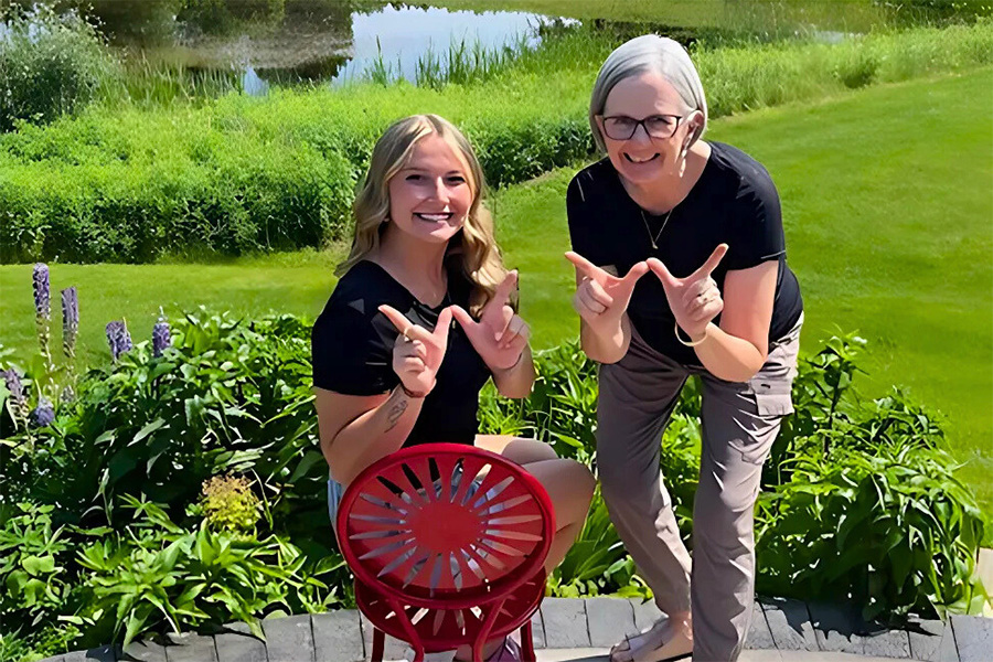 Gracie DeKeyser and Emily Mroczynski make the W hand sign while posing in front of a pond surrounded by bright green grass.