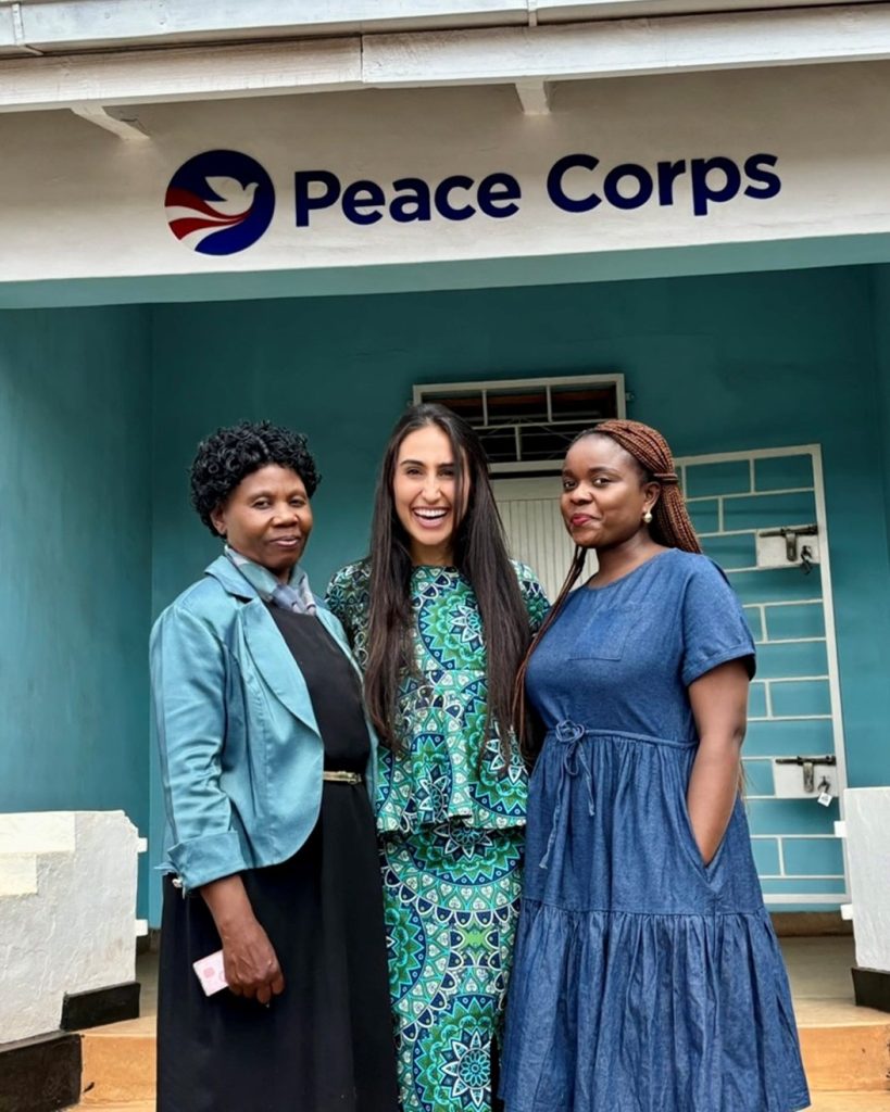 Gonzalez stands with two Peace Corps colleagues in Malawi, under a sign with the Peace Corps emblem.