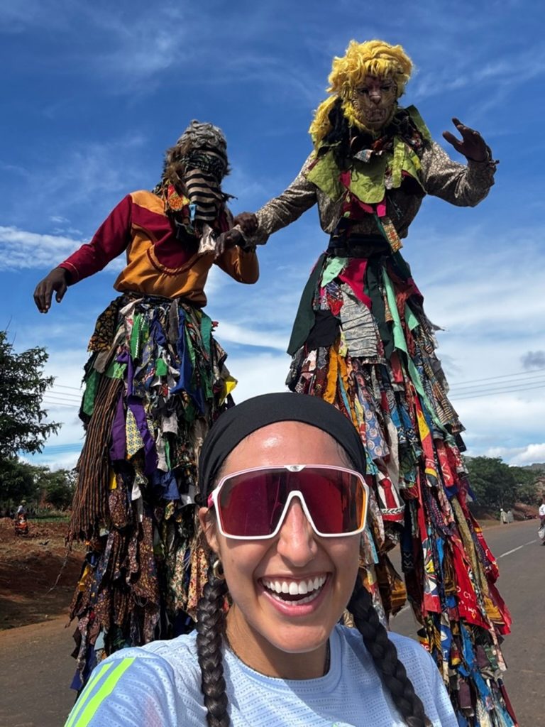 Gonzalez smiling as two Malawian stilt walkers in traditional costumes stand behind her.
