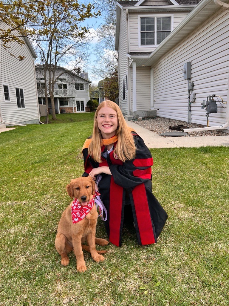 Rachel Stanek kneels in graduation robes with her golden retriever puppy.