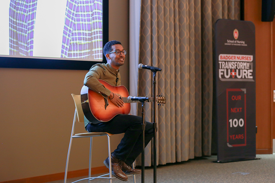 Tad Worku, speaker for the Littlefield Leadership Lecture, wears a brown shirt and strums a guitar while sitting next to a microphone. He is singing.