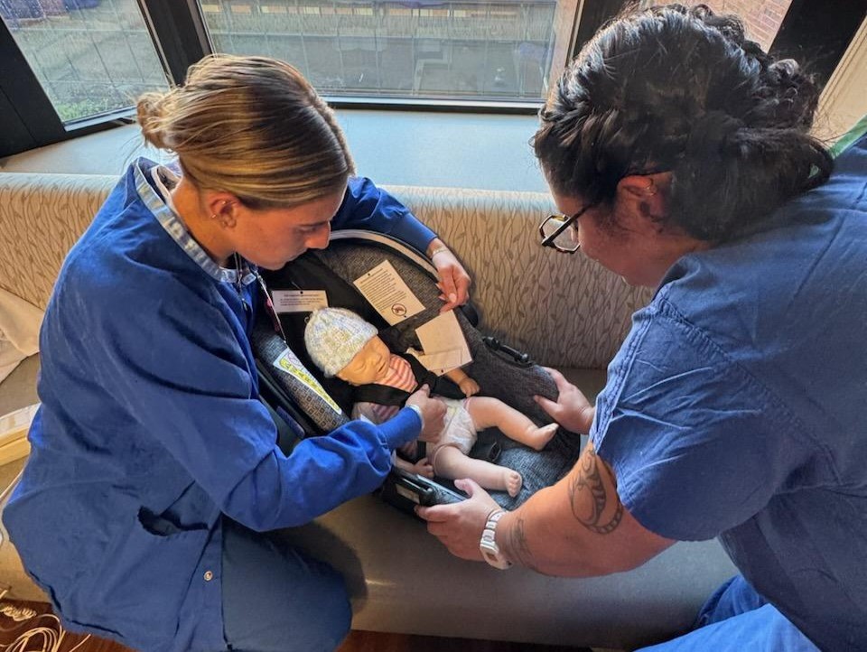 Nursing students in blue scrubs strap an infant medical mannequin into a car seat.