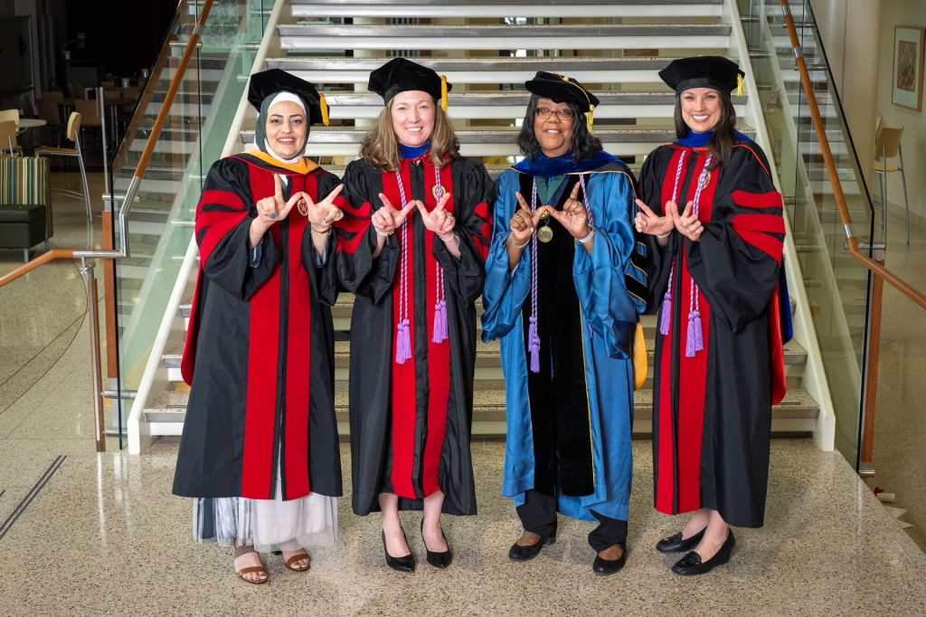 Abby Hammes and Dean Emerit Linda Scott with two other PhD graduates, all in caps and gowns. They stand at the foot of the stairs in Cooper Hall making the W hand sign.