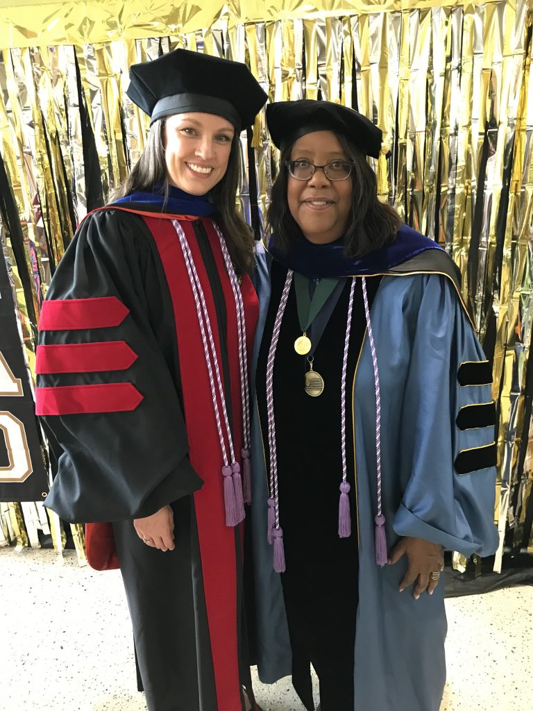 Jessica LeClair and Dean Emerit Scott pose for a picture in their full graduation regalia against a background of gold streamers.