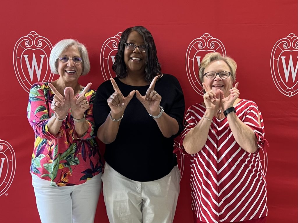 Linda Hurwitz and Linda Scott stand against a red UW photo background and make the W hand sign with another colleague.