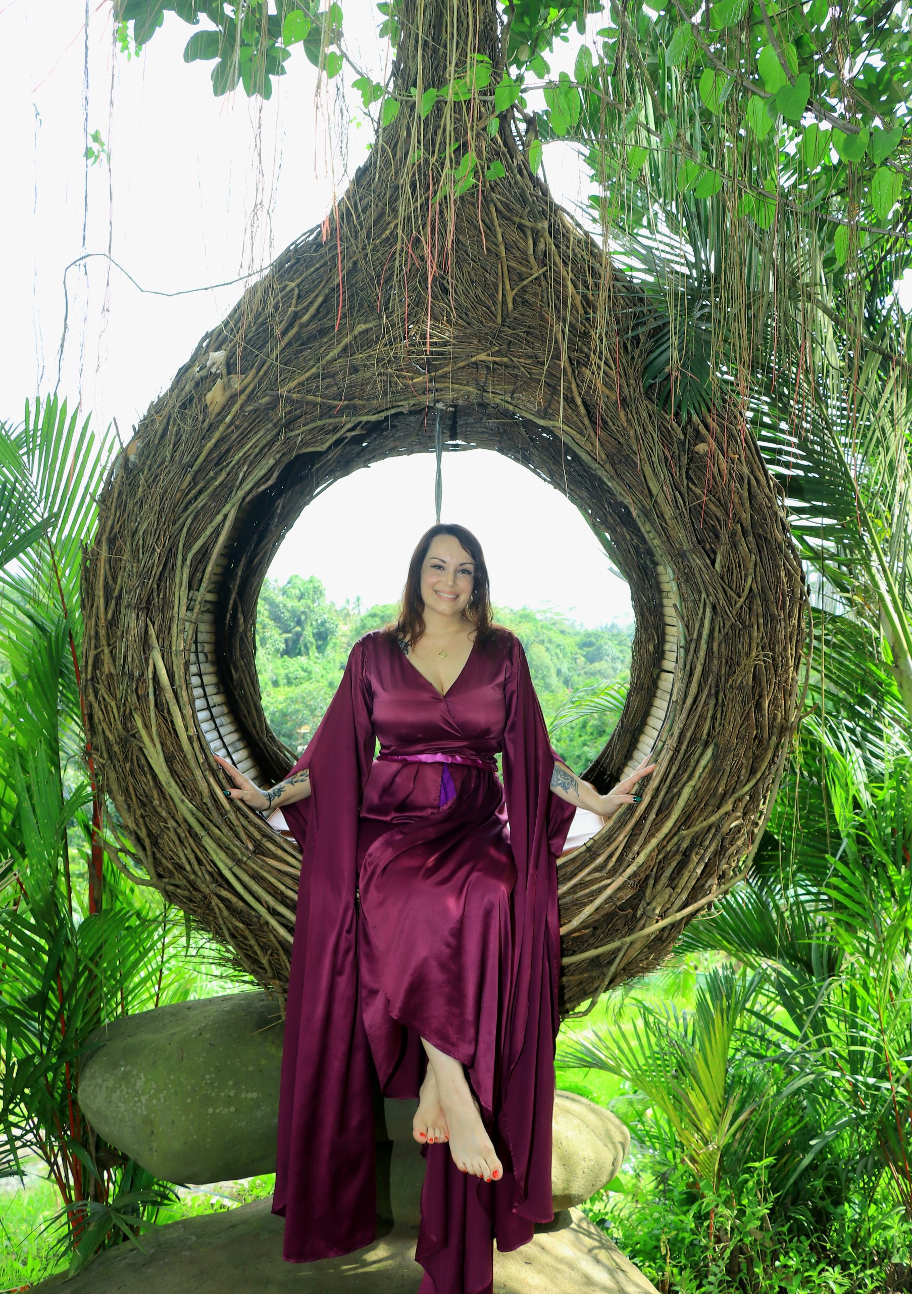 Henze wears a flowing burgundy dress while sitting inside a round swing made of branches on a recent vacation.