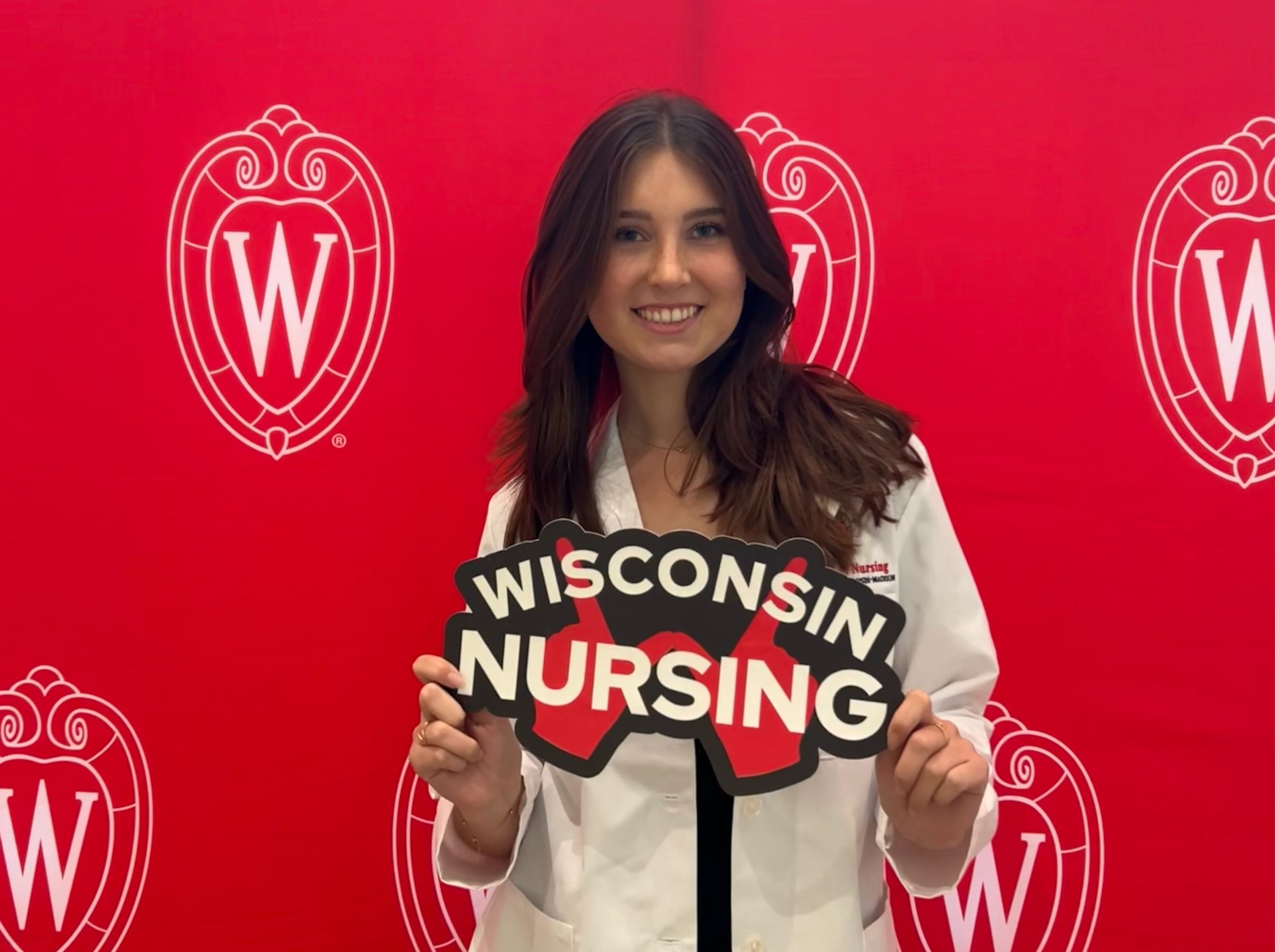 Taylor Brook wears a white coat and stands against a red background with the UW crest. She holds a sign that says Wisconsin Nursing.