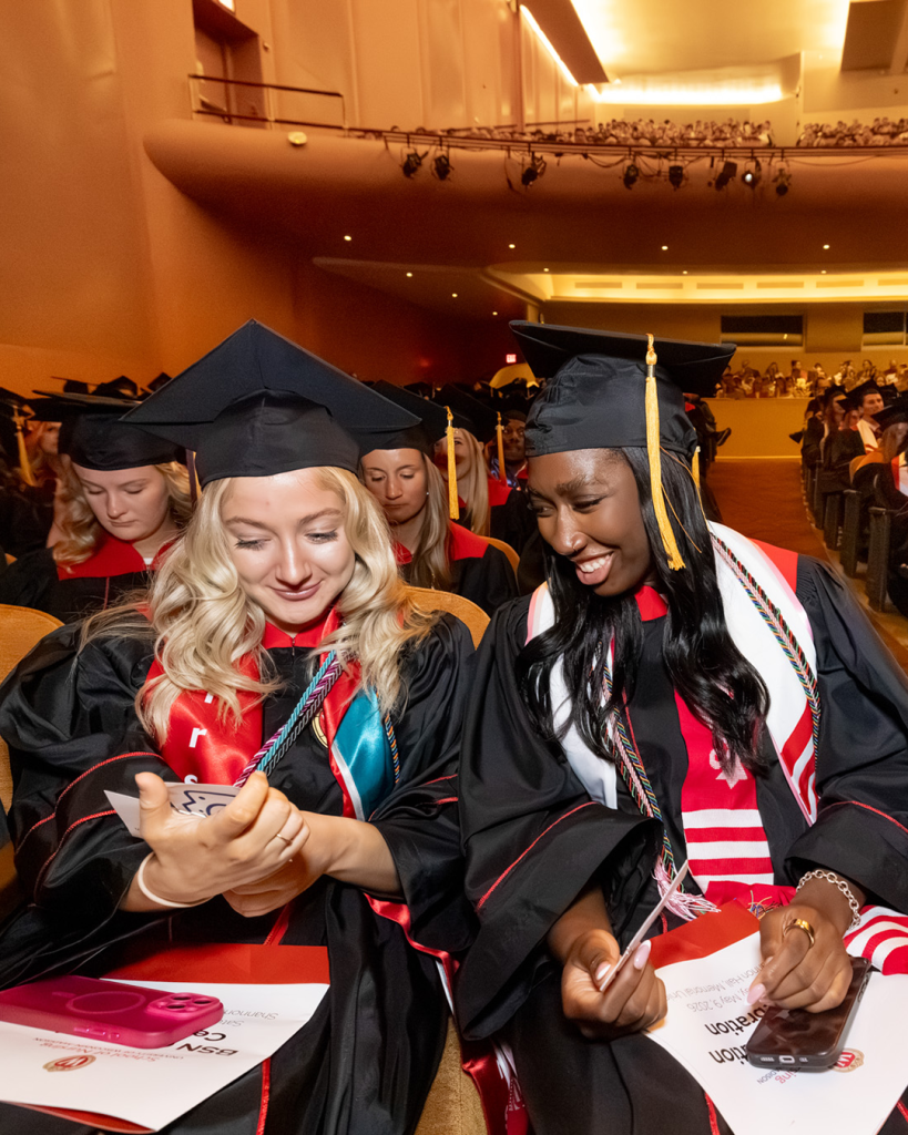 Macy Deaton and a fellow BSN student sit next to each other in their caps and gowns at graduation, smiling as they look at the program.