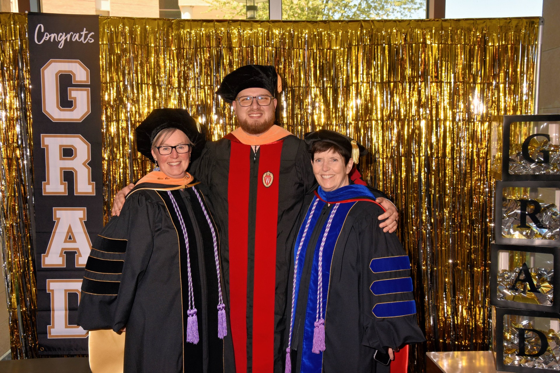Joshua Hermsdorf in cap and gown standing in front of a glittery gold background with his arms around two faculty members from his DNP program.