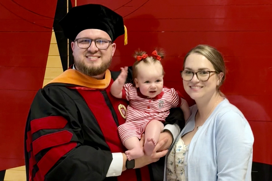 Hermsdorf in cap and gown stands next to his wife while holding his smiling baby daughter.