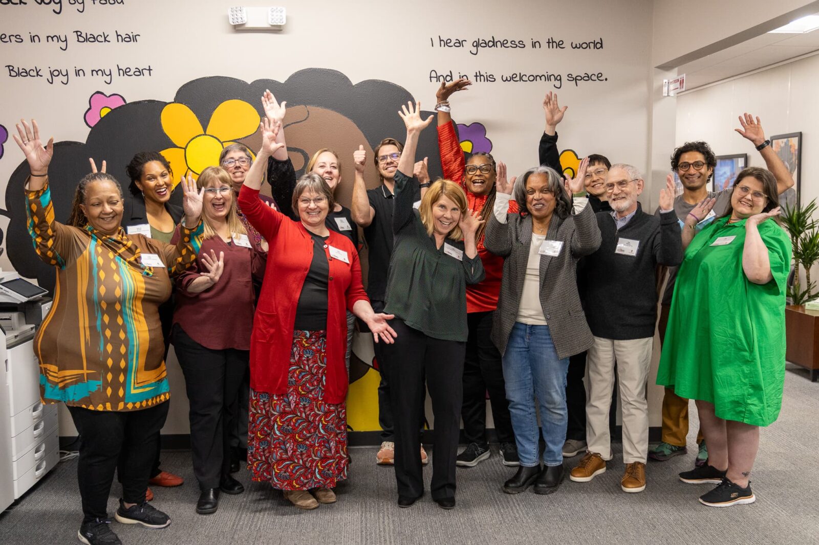 School of nursing faculty and The Community-Academic Aging Research Network (CAARN) participants smile and throw their hands in the air in front of a colorful mural. Photo credit: Dynae Allice.